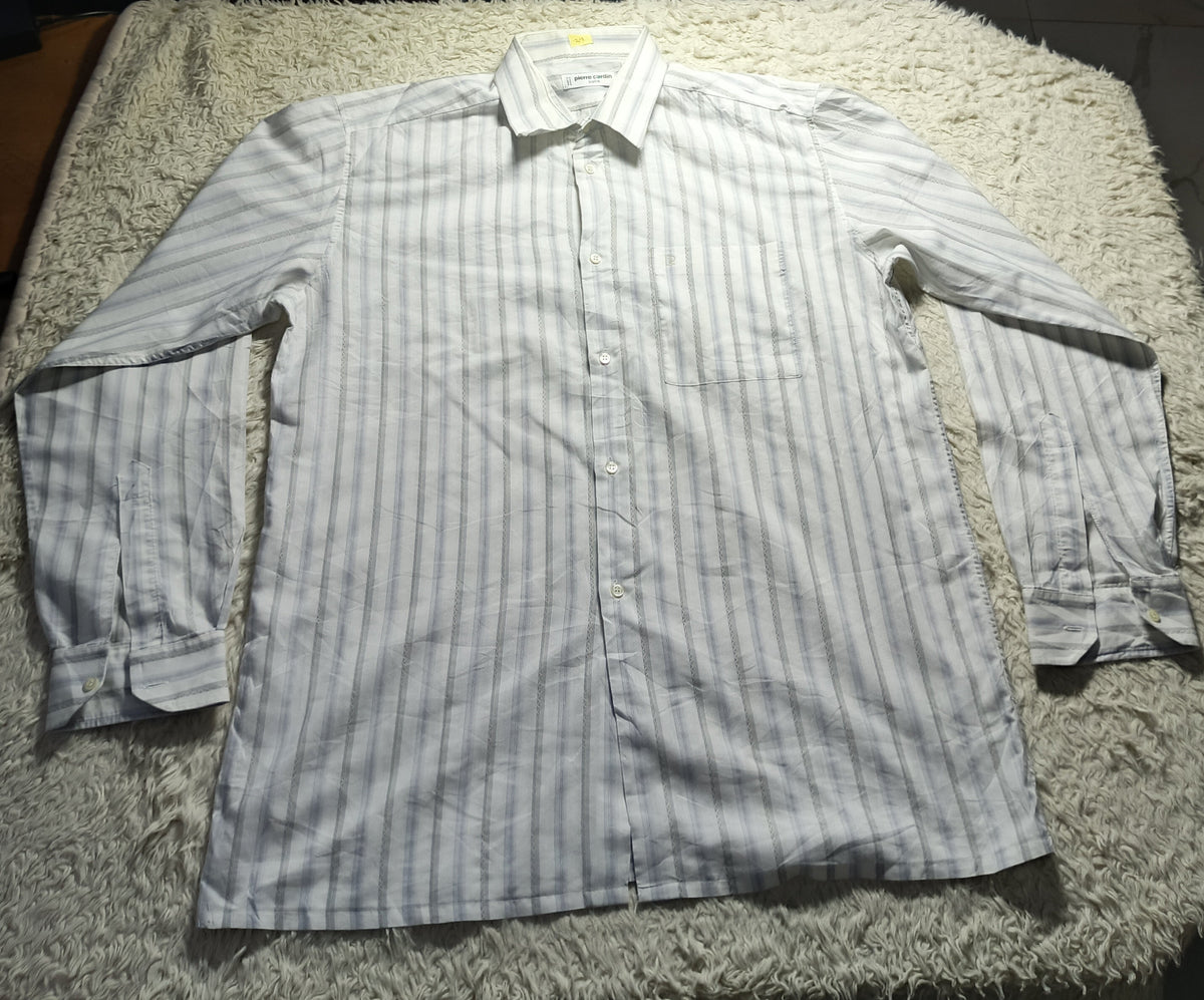 A long-sleeve button-up shirt with thin vertical stripes in light gray and white, laid flat on a shaggy cream rug. The photo is taken in a dimly lit room with wooden chairs and a light stand visible in the background.