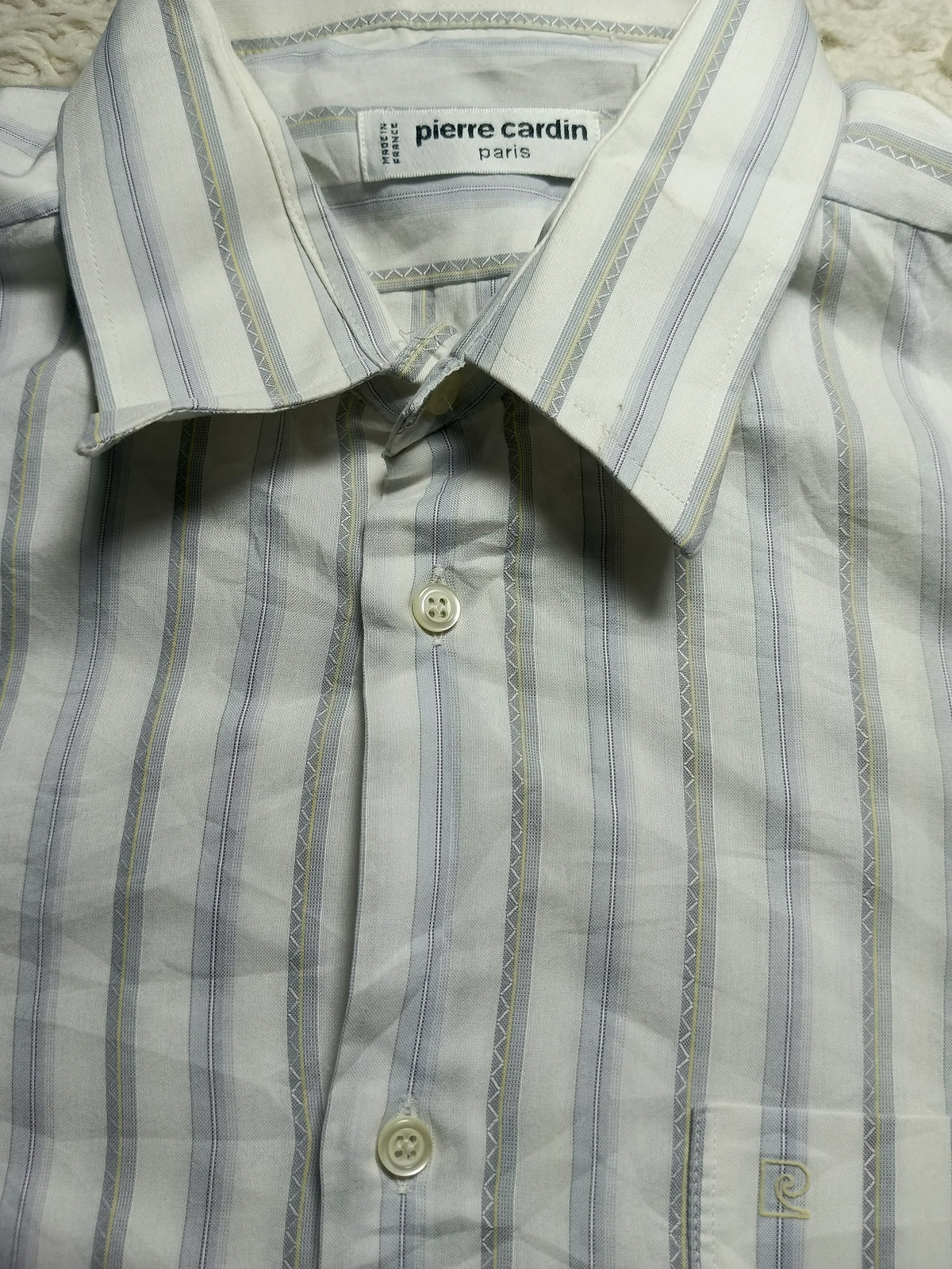 A close-up of a Pierre Cardin Paris button-down shirt featuring a white base with vertical light blue and beige stripes, laid flat on a textured white fabric background.