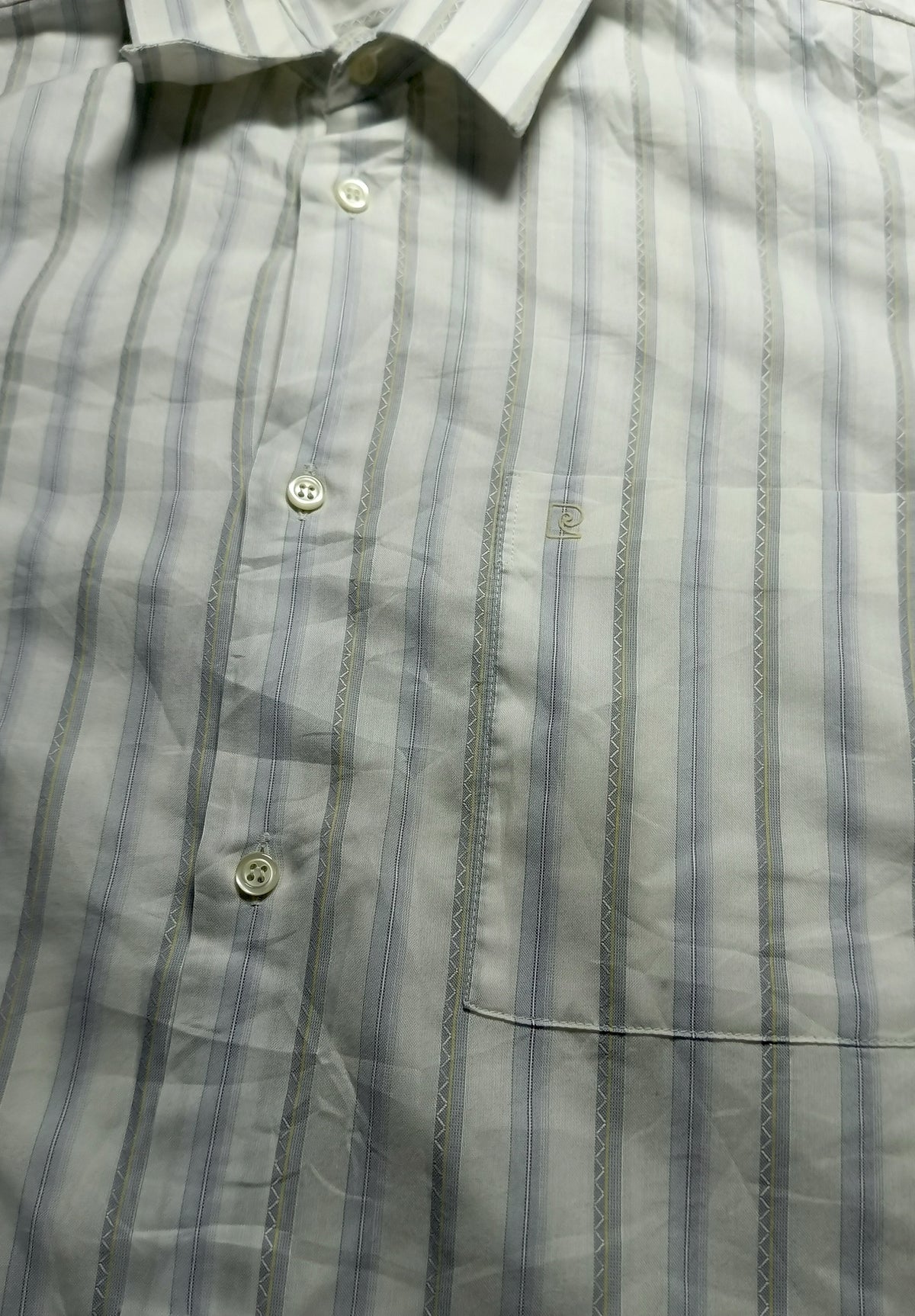 A close-up of a button-down shirt with vertical stripes in light blue, gray, and white, featuring a collar, three visible buttons, and a chest pocket.