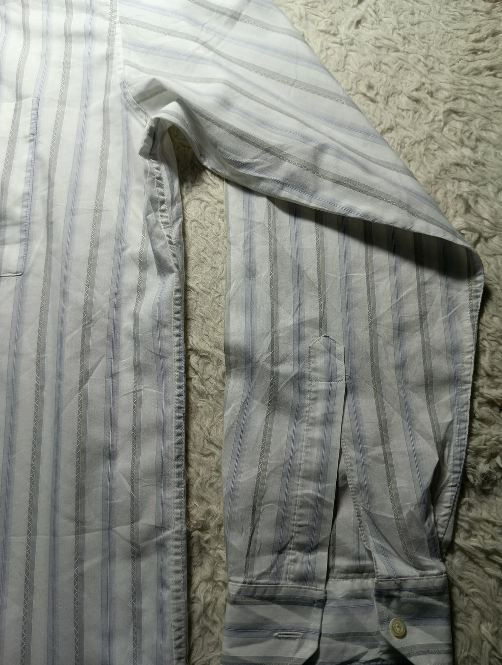 A close-up of a light gray and white vertically striped long-sleeve shirt laid flat on a textured beige rug. The photo is angled, showing the chest pocket, sleeve, and buttoned cuff.