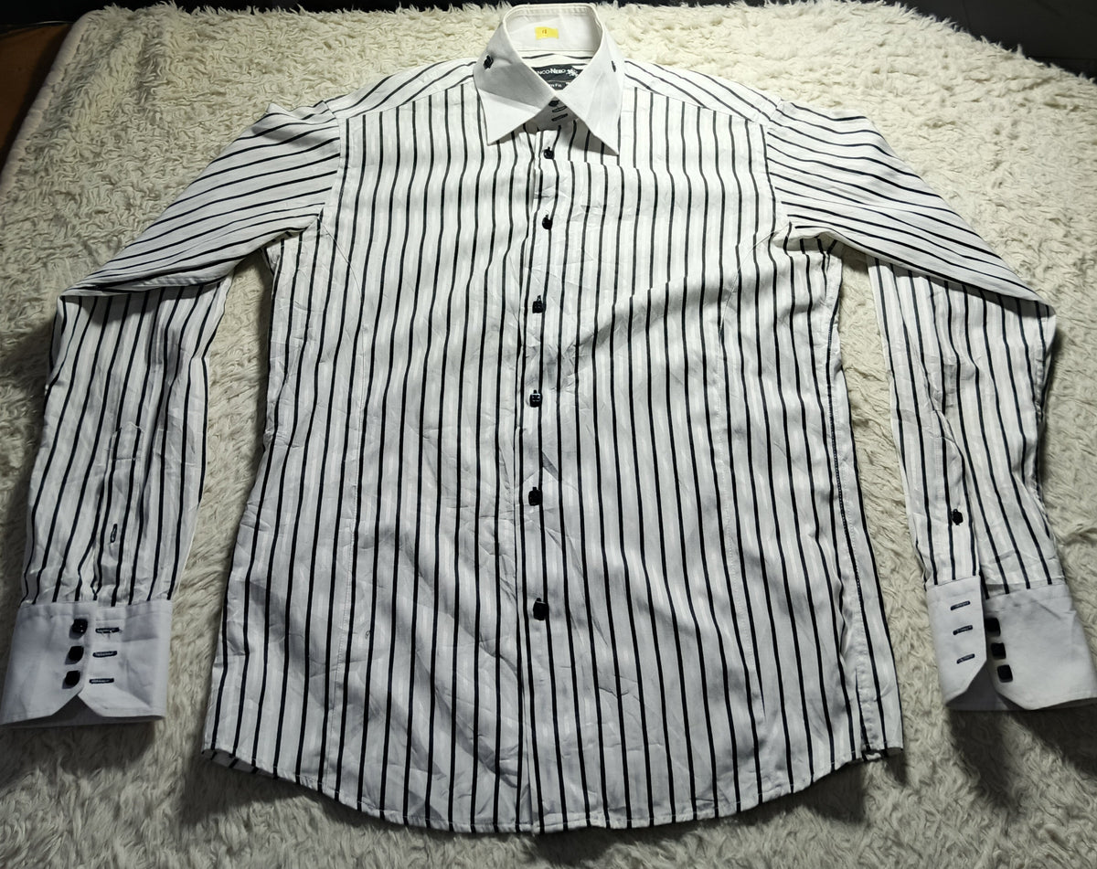 A long-sleeve button-up dress shirt with thin black vertical pinstripes on a white base, laid flat on a white shag rug. A small LED light is shining onto the shirt from above, with chairs and a red garment visible in the background.
