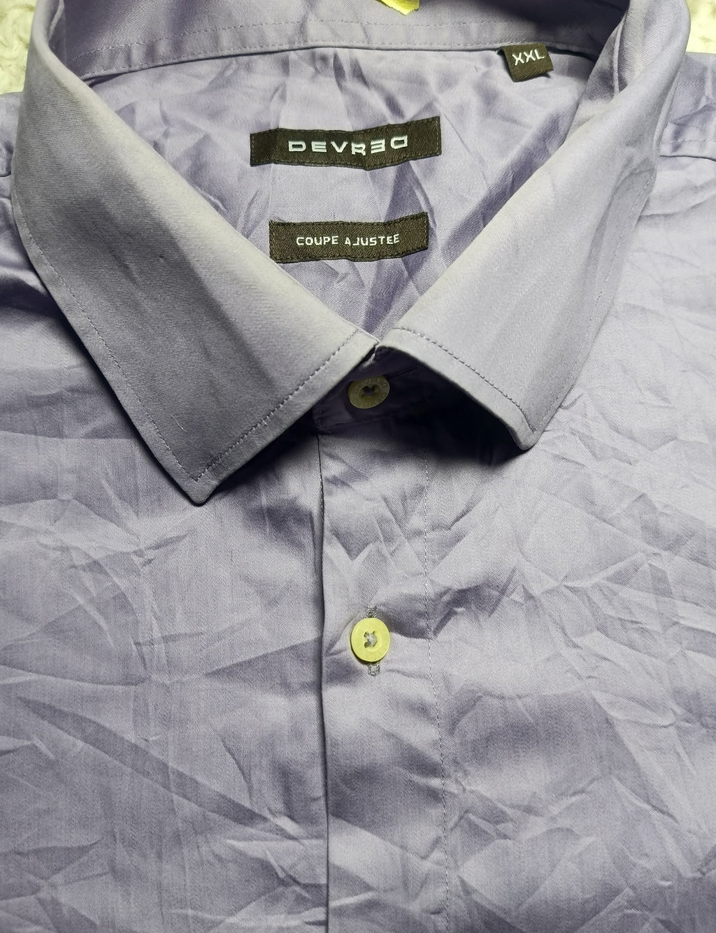 A close-up of a light blue-gray collared shirt by Deverso, showing the inner neck label, collar, and front button. The shirt has a crinkled texture and is photographed flat against a neutral background.