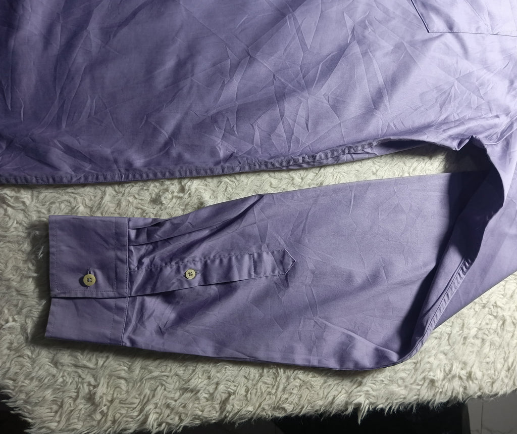 A lavender purple long-sleeve button-up shirt laid flat on a white textured rug, showing the fabric texture, collar, and buttoned cuff.