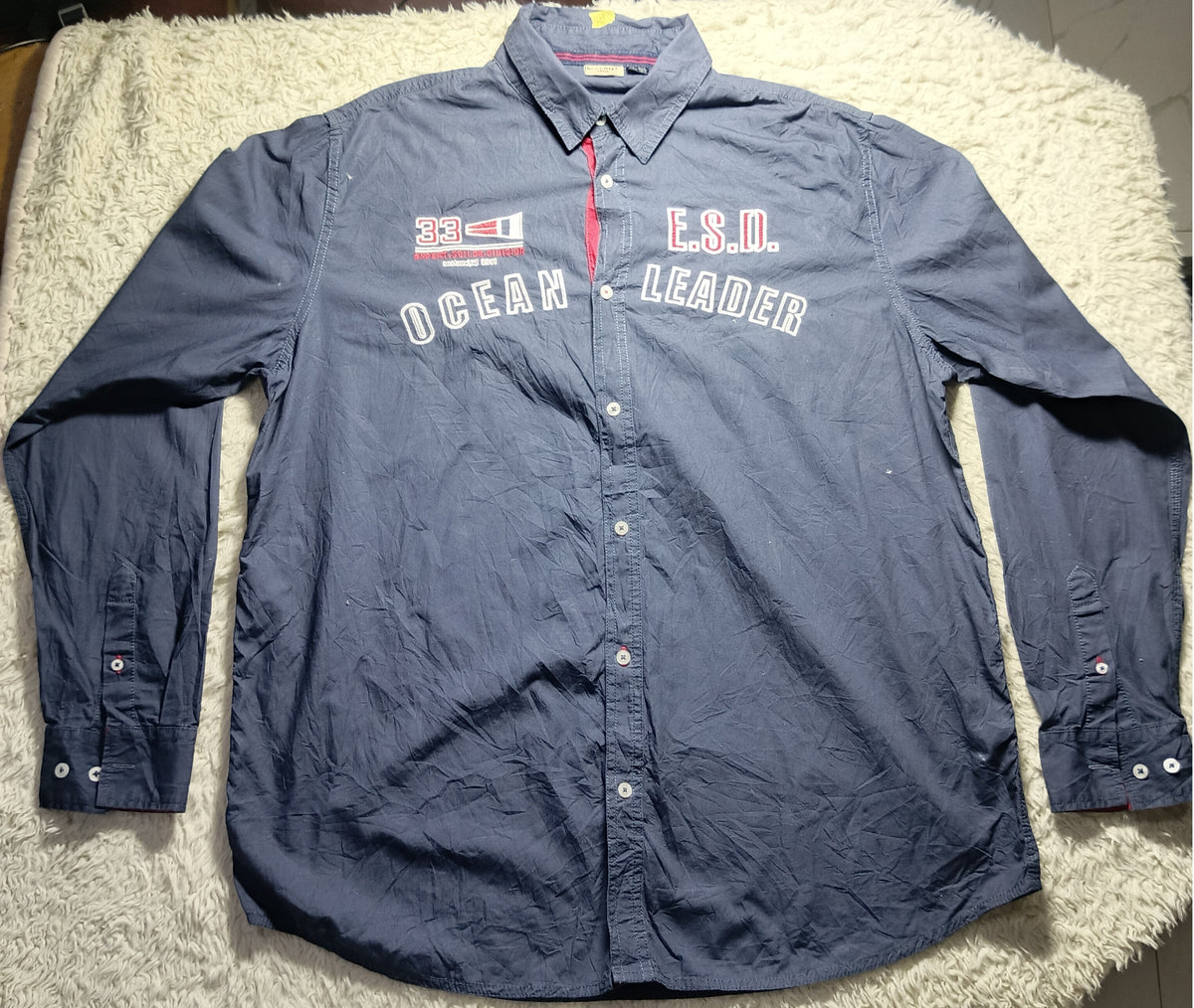 A navy blue long-sleeve button-up shirt laid flat on a white shag rug. The shirt features 'SEMPER FALSA' and 'DESIGN FALSA' printed in white and red on the left chest.