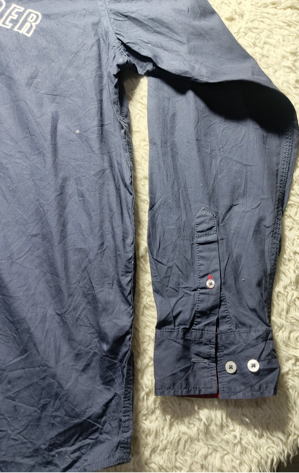 A close-up of a dark navy blue long-sleeve shirt laid flat on a white shaggy rug, showing the sleeve, cuff with buttons, and part of the torso.