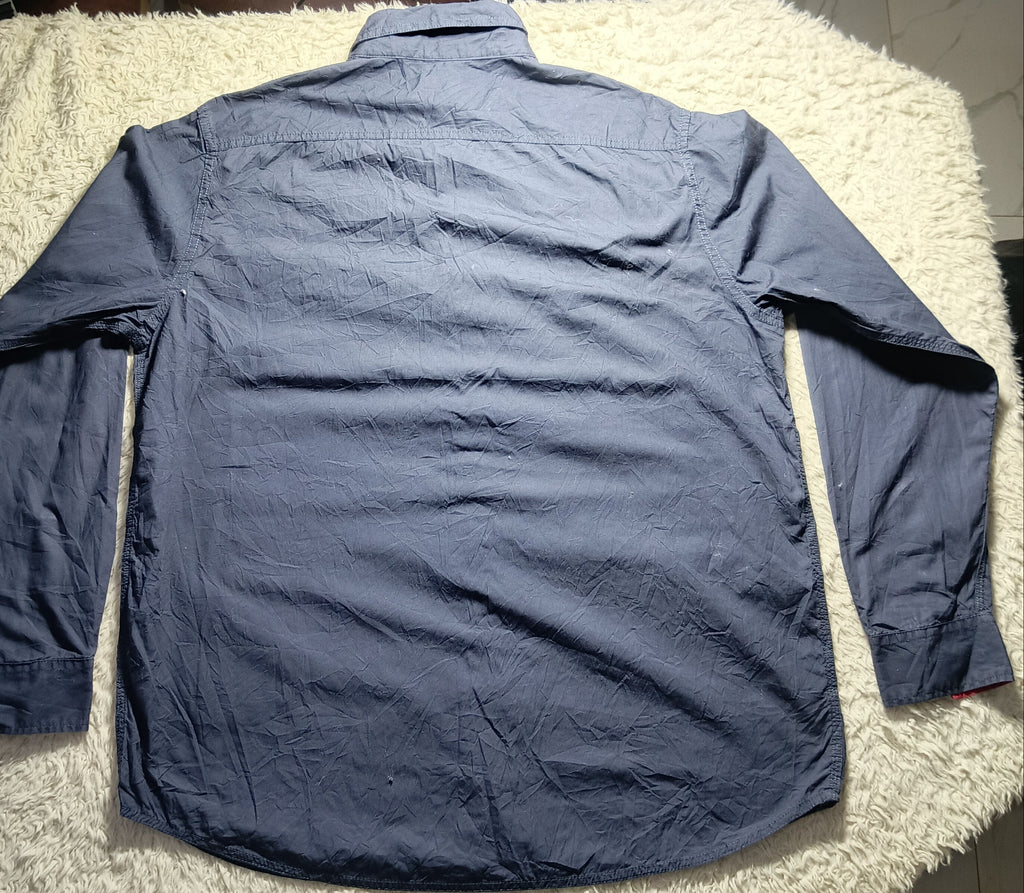 A long-sleeve blue button-up shirt laid flat on a white shag rug, photographed from above in an indoor setting with a tiled floor and bags in the background.