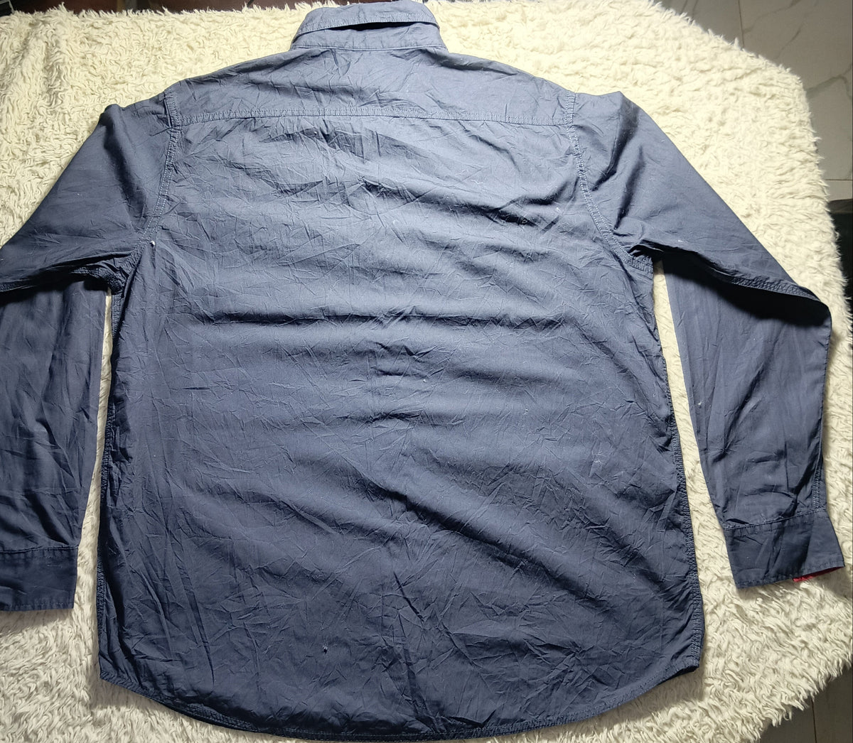 A long-sleeve blue button-up shirt laid flat on a white shag rug, photographed from above in an indoor setting with a tiled floor and bags in the background.