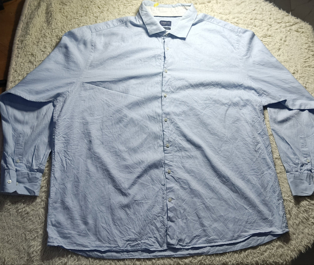 A light blue long-sleeve button-up shirt laid flat on a beige shaggy blanket, photographed indoors with a dark chair and table visible in the background.