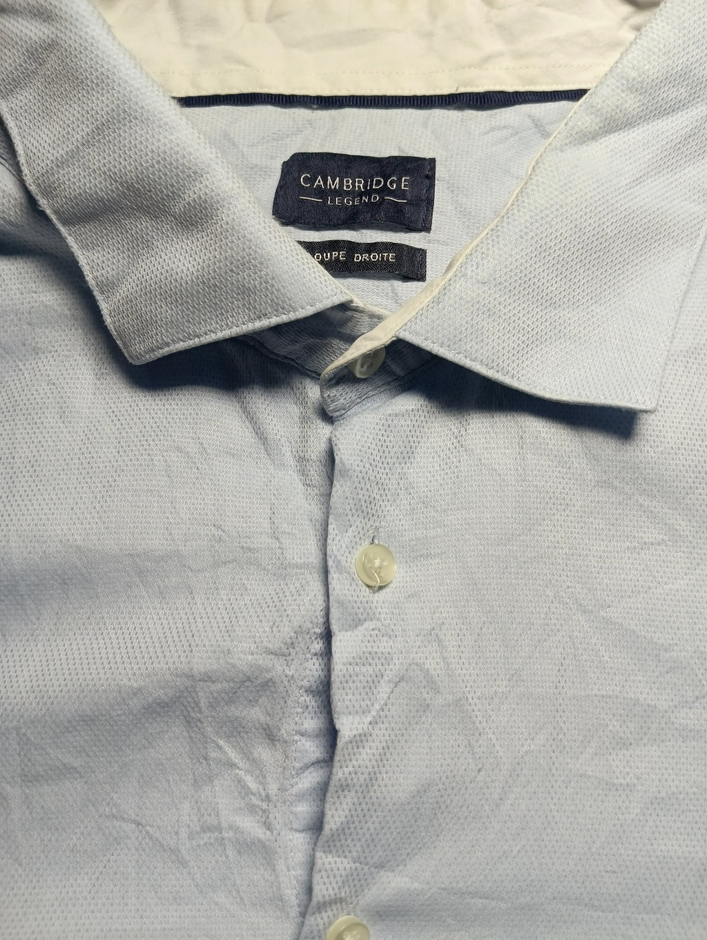 Close-up of a light blue textured Oxford shirt collar with a visible Cambridge University Press label, showing the fabric weave and buttons.