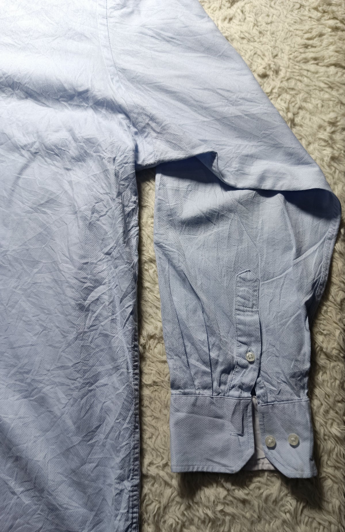 A light blue long-sleeve button-up shirt laid flat on a textured beige rug, with the right sleeve folded back to show the cuff and button details.