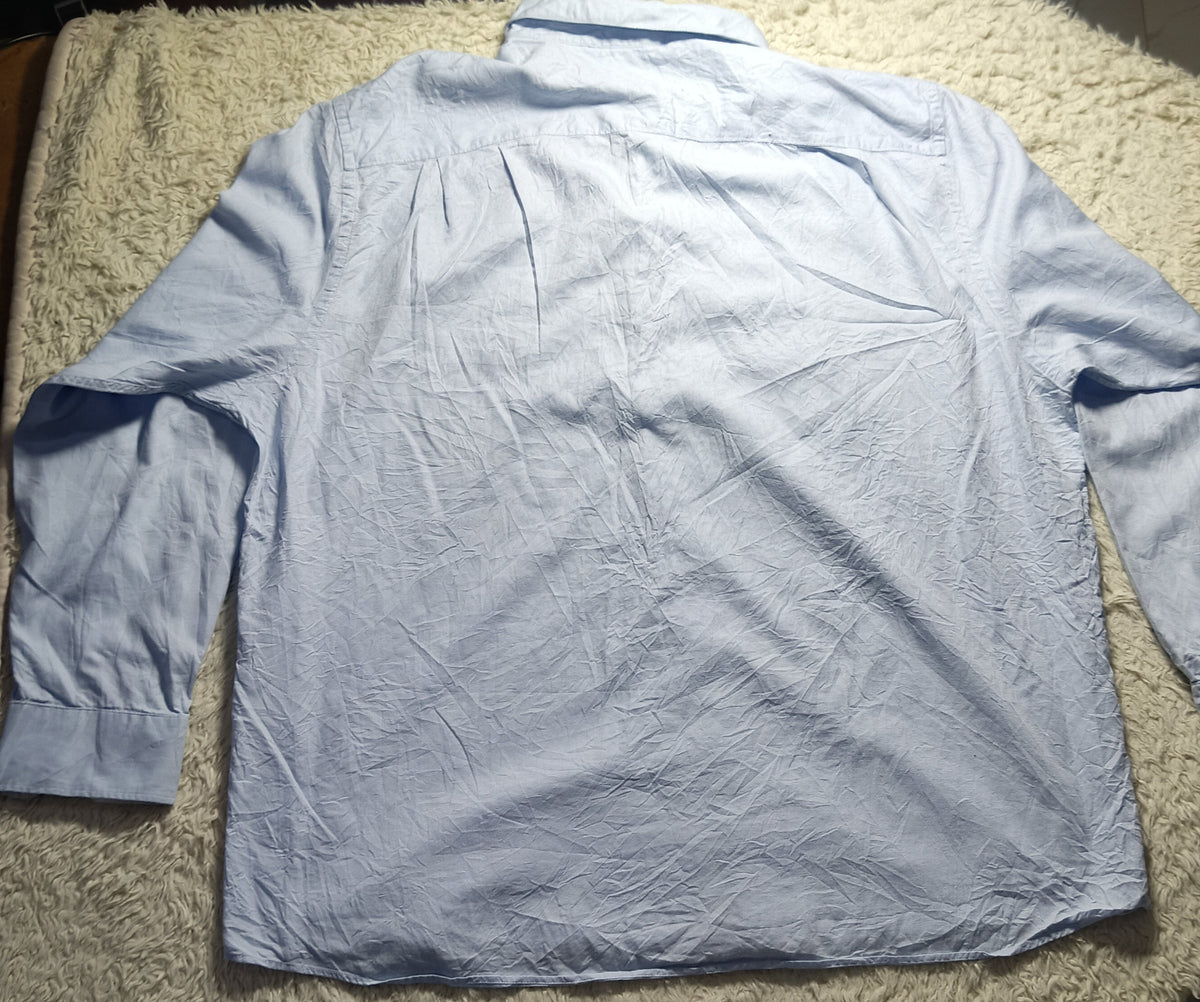 A light blue long-sleeve button-up shirt laid flat on a beige shaggy rug, photographed from above in an indoor setting with tiled floor visible.