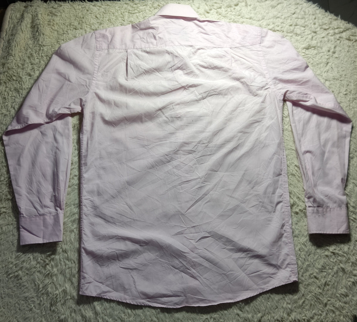 A light pink long-sleeve shirt laid flat on a textured white shag rug, photographed from above with visible wrinkles.