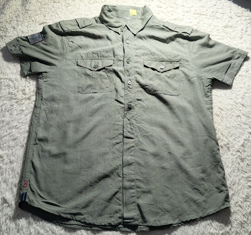 A short-sleeve button-up shirt in a muted olive green color, laid flat on a white shag rug. The shirt features two buttoned chest pockets and a small patch on the right sleeve.