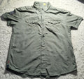 A short-sleeve button-up shirt in a muted olive green color, laid flat on a white shag rug. The shirt features two buttoned chest pockets and a small patch on the right sleeve.