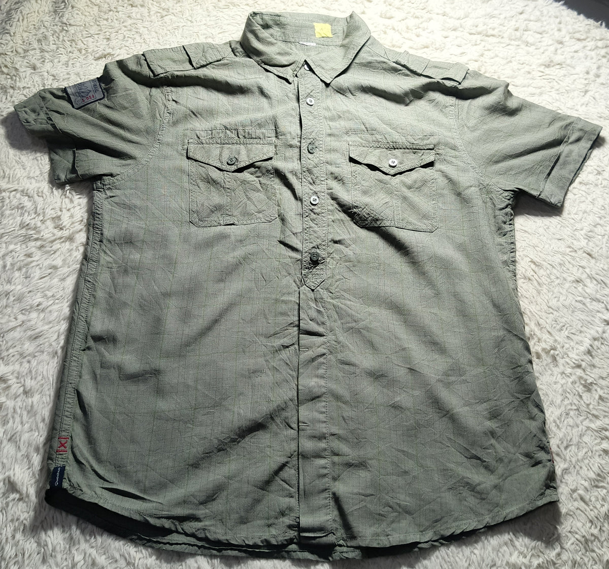 A short-sleeve button-up shirt in a muted olive green color, laid flat on a white shag rug. The shirt features two buttoned chest pockets and a small patch on the right sleeve.