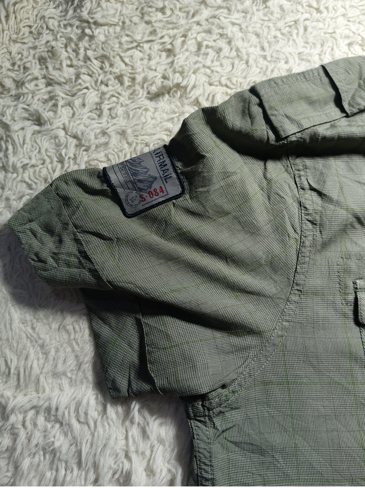 An olive green long-sleeve shirt laid flat on a white shag rug, showing the inner collar with a brand label that reads 'SOUZA'.