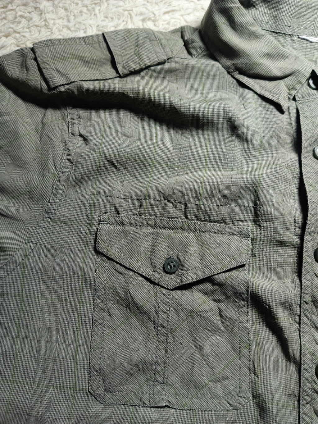 A close-up of a dark olive green textured button-up shirt with a chest pocket, laid flat on a light patterned surface.