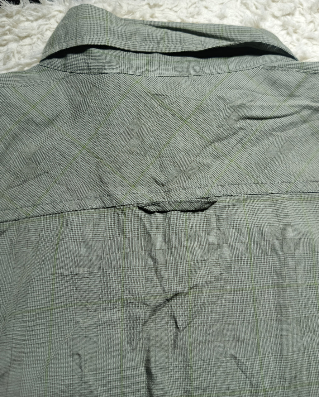 Close-up of the back of a wrinkled olive green plaid shirt laid flat on a white shag rug, with camera metadata text at the bottom.
