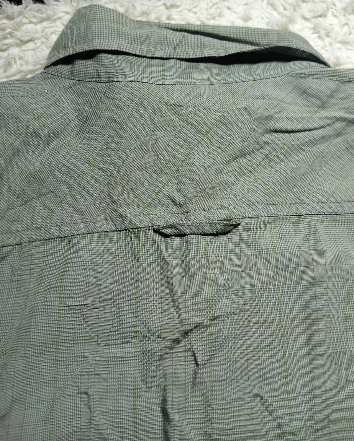 Close-up of the back of a wrinkled olive green plaid shirt laid flat on a white shag rug, with camera metadata text at the bottom.