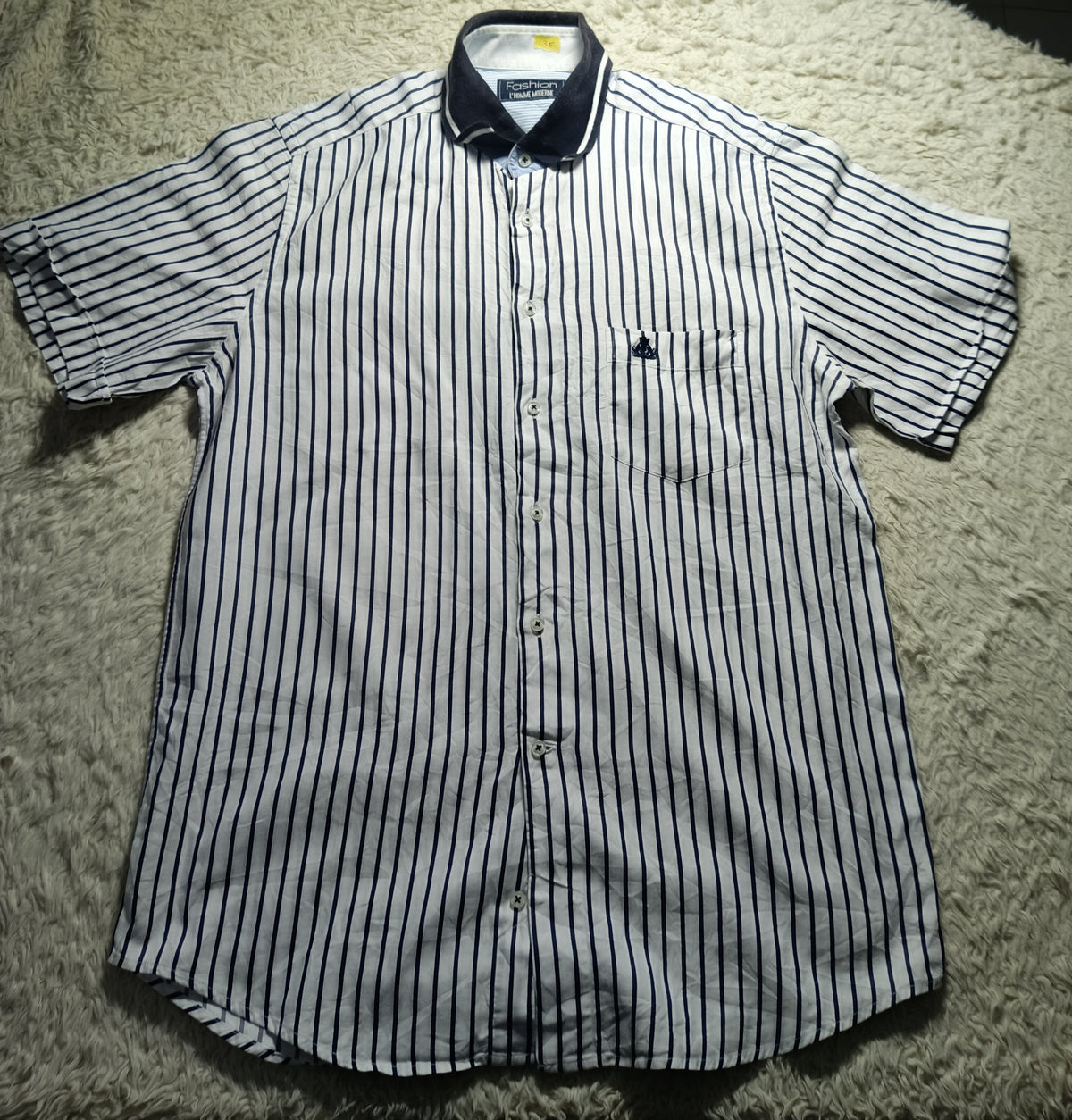 A short-sleeve button-up shirt with thin vertical navy blue stripes on a white background, laid flat on a white shag rug. The shirt features a dark navy collar and a small embroidered logo on the left chest pocket.