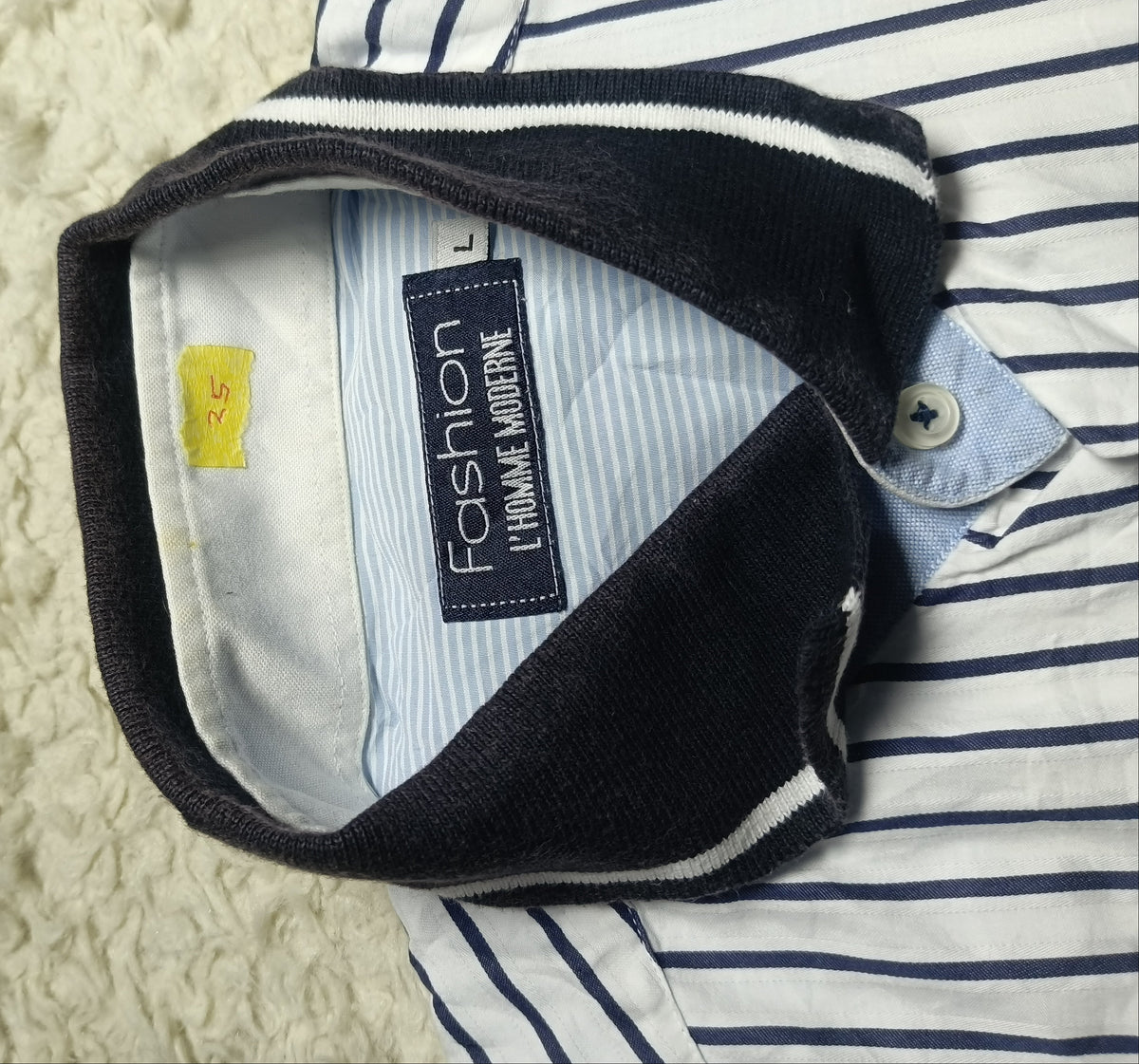 Close-up of a striped button-up shirt laid flat on a white textured blanket, with the collar turned up to show the inside neck label reading 'Fashion 1 Volume Worldwide'.