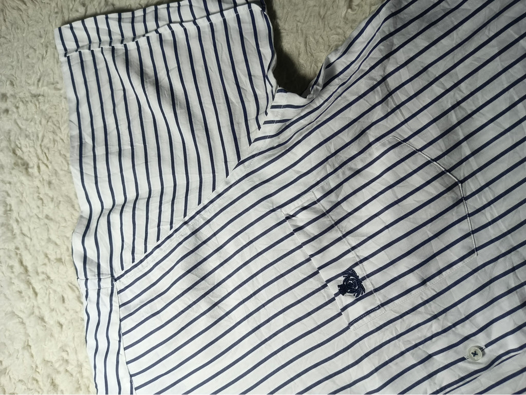 A close-up of a white short-sleeve shirt with thin navy blue vertical stripes, laid flat on a textured cream-colored rug. A small embroidered logo is visible on the chest pocket area.