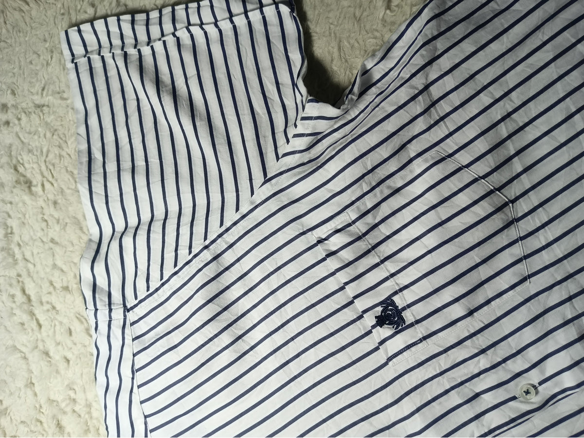 A close-up of a white short-sleeve shirt with thin navy blue vertical stripes, laid flat on a textured cream-colored rug. A small embroidered logo is visible on the chest pocket area.