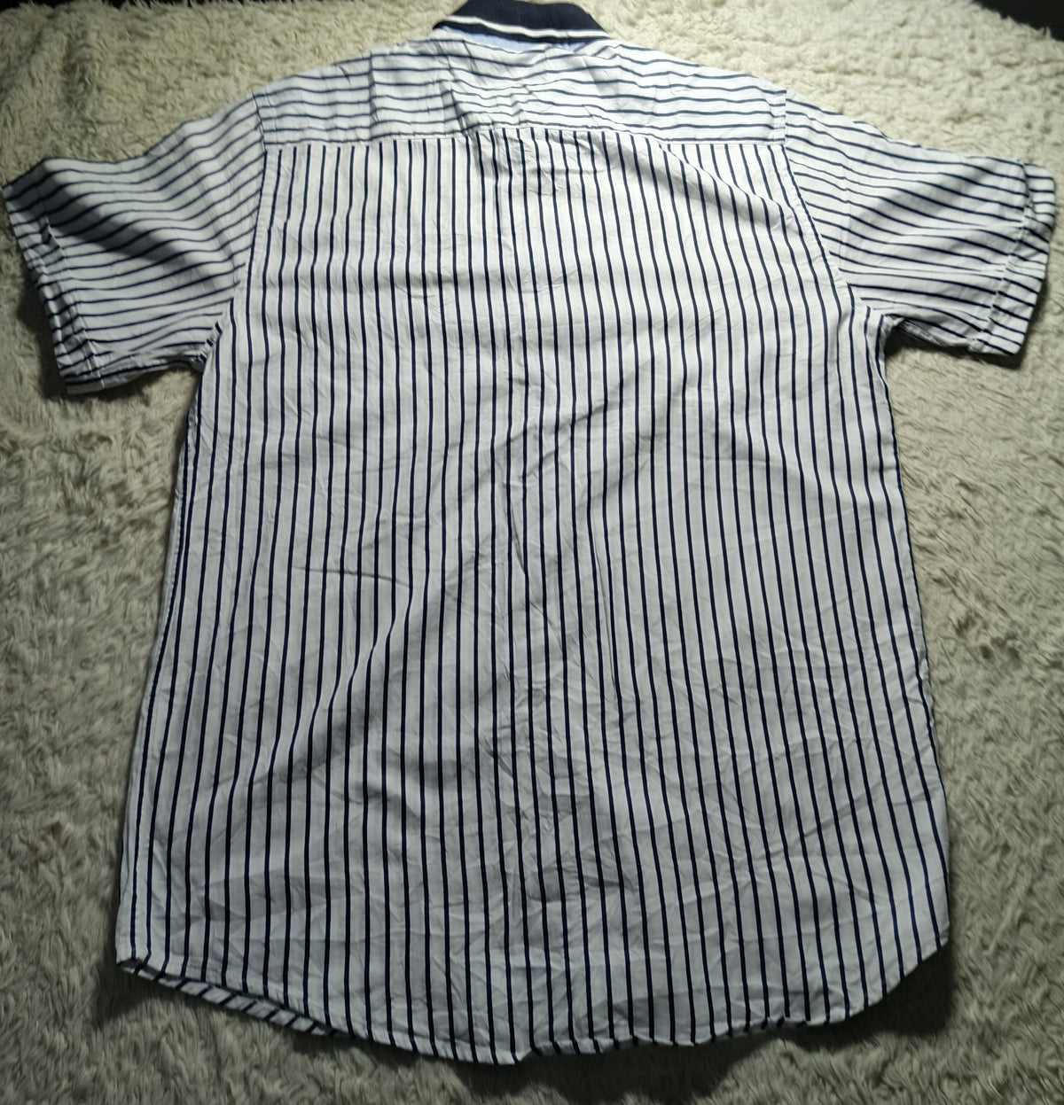 A short-sleeve button-up shirt with thin vertical navy blue stripes on a white or off-white base, laid flat on a white shag rug.