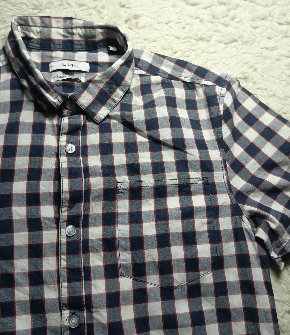 A short-sleeve button-up plaid shirt laid flat on a white textured rug. The shirt features a navy blue, white, and thin red checkered pattern, with a single chest pocket and visible 'A.