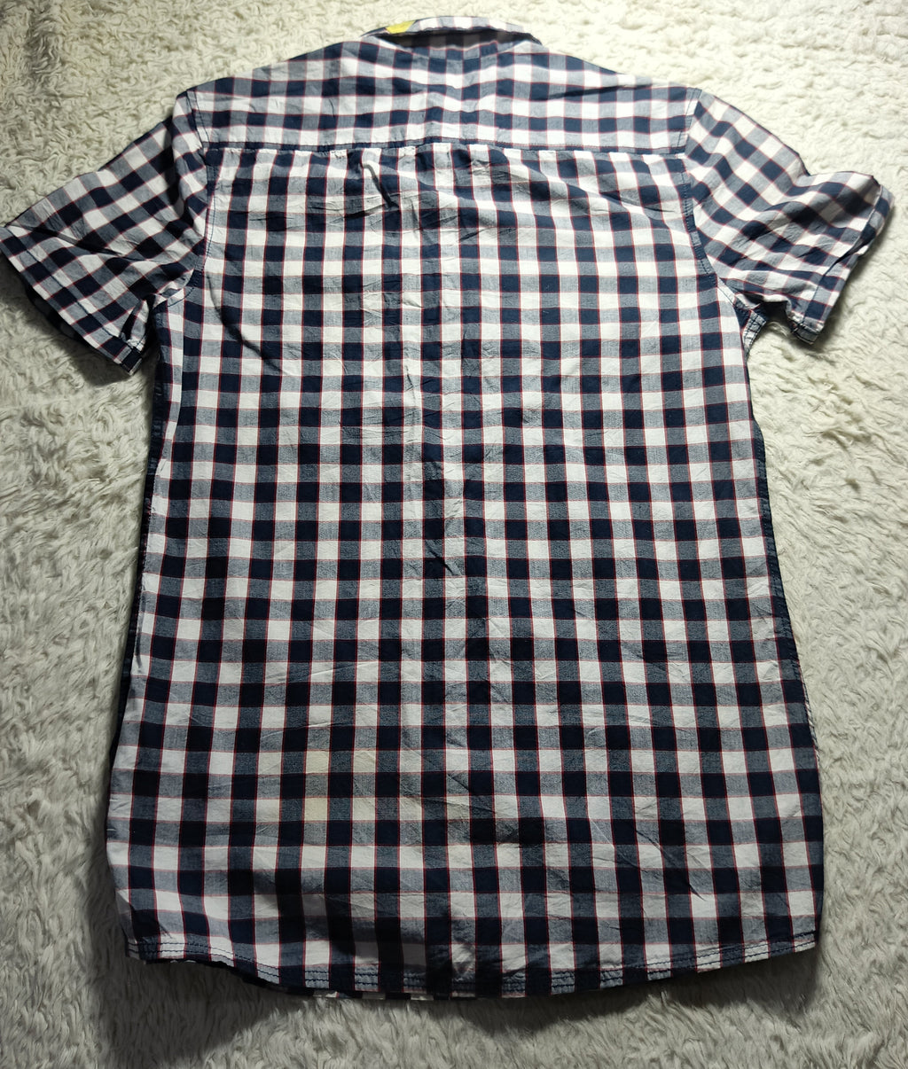 A short-sleeve button-up shirt with a navy blue, white, and light gray checkered pattern, laid flat on a white shag rug.