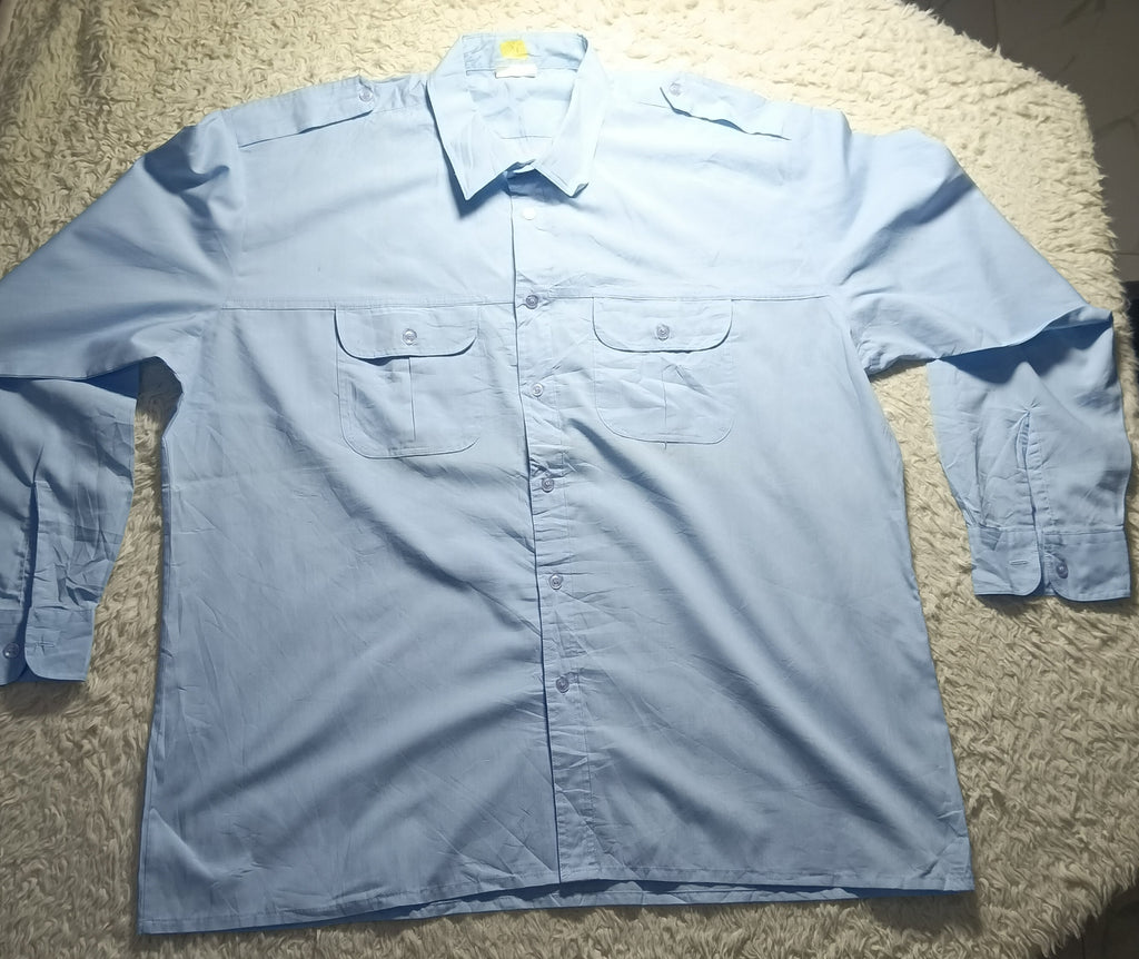 A light blue long-sleeve button-up shirt with two chest pockets, laid flat on a white shaggy blanket in a home setting.