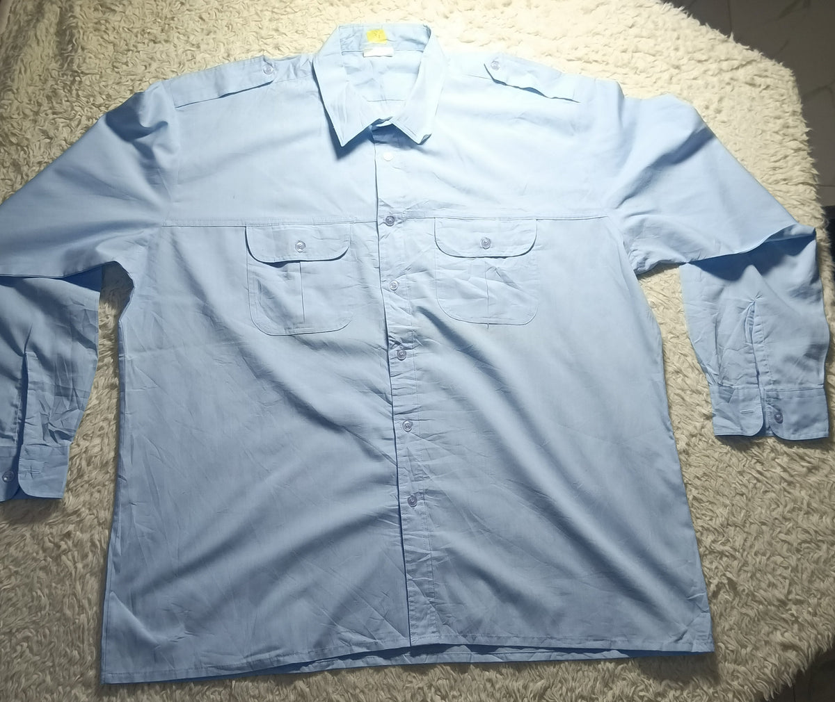 A light blue long-sleeve button-up shirt with two chest pockets, laid flat on a white shaggy blanket in a home setting.