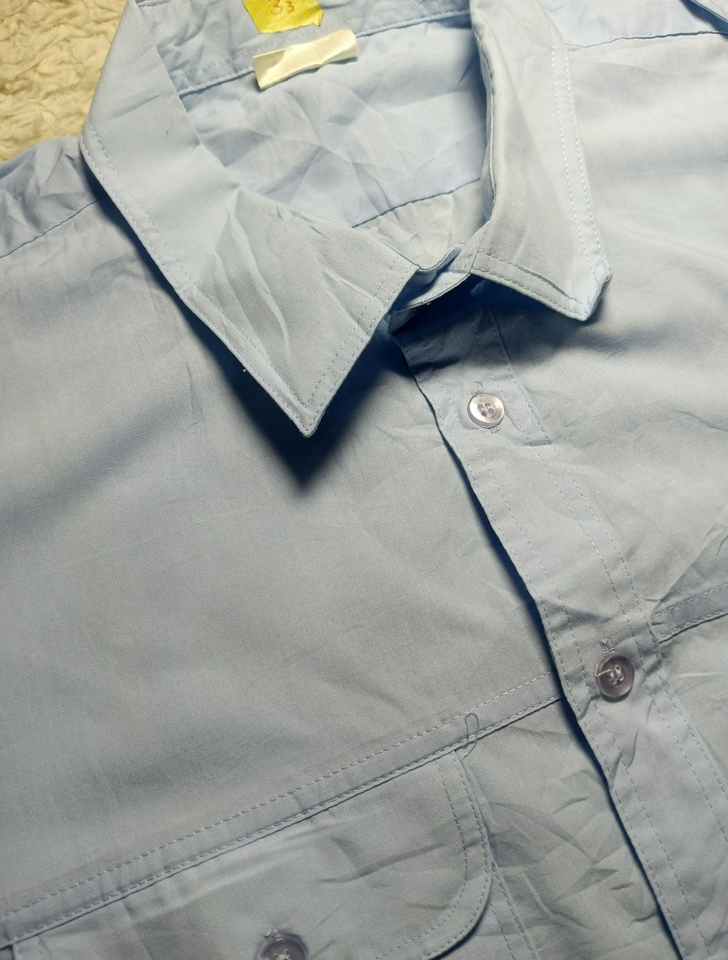 A light blue button-down shirt laid flat, showing the collar, front buttons, and part of a chest pocket. The photo is taken close up on a textured beige surface, with a camera watermark at the bottom.