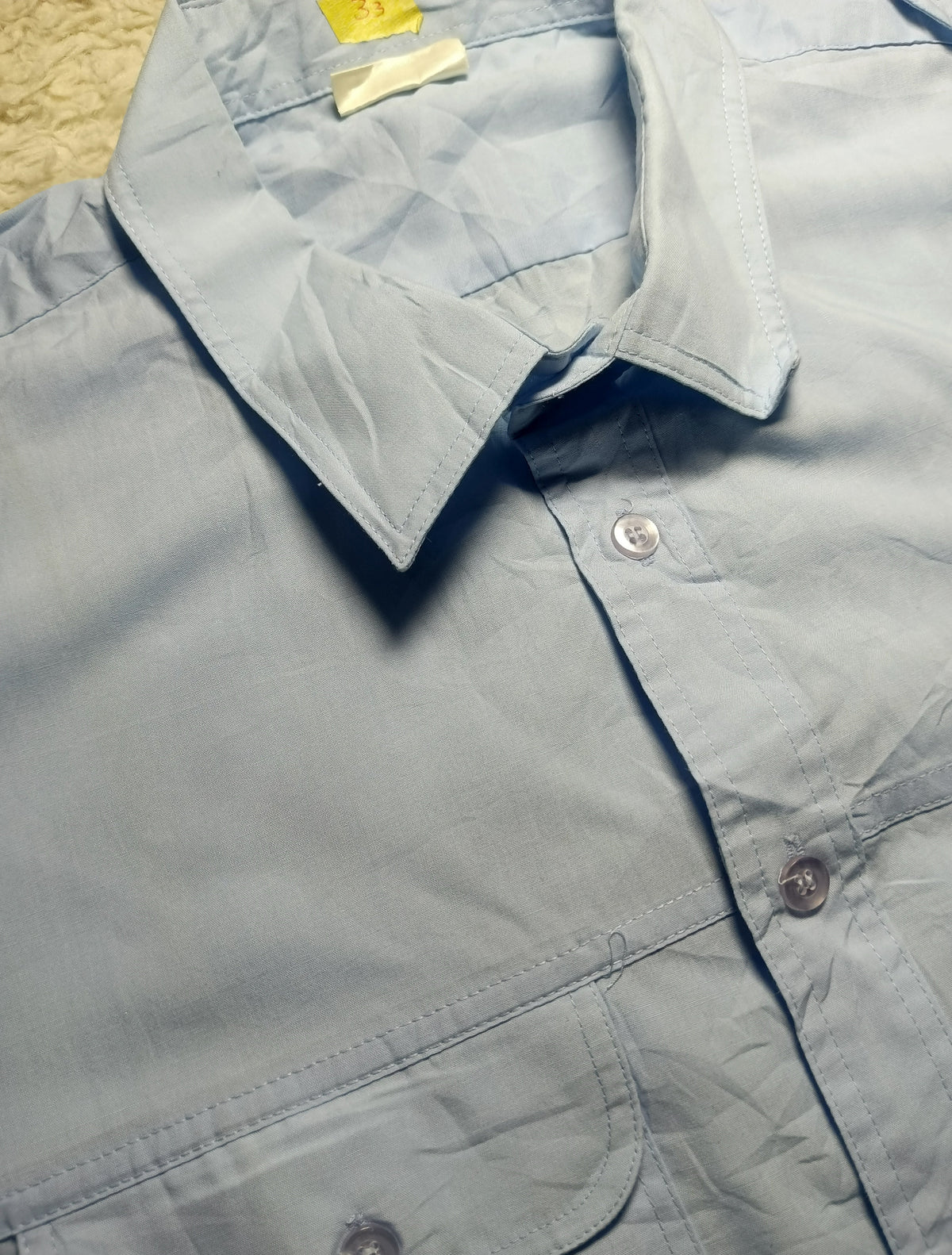 A light blue button-down shirt laid flat, showing the collar, front buttons, and part of a chest pocket. The photo is taken close up on a textured beige surface, with a camera watermark at the bottom.