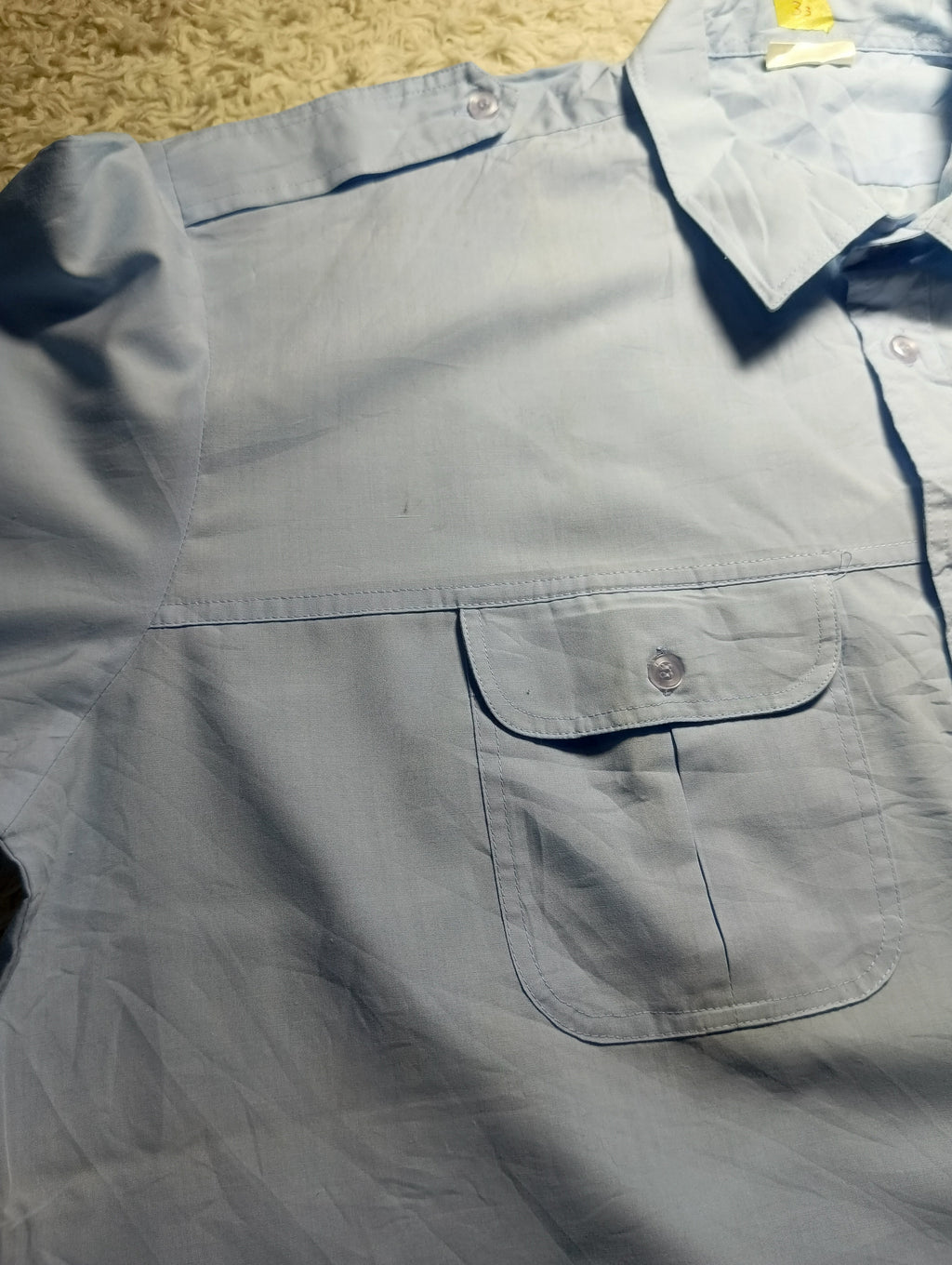 A light blue long-sleeve button-up shirt laid flat on a textured beige carpet. The shirt features a chest pocket with a button flap and a collar.