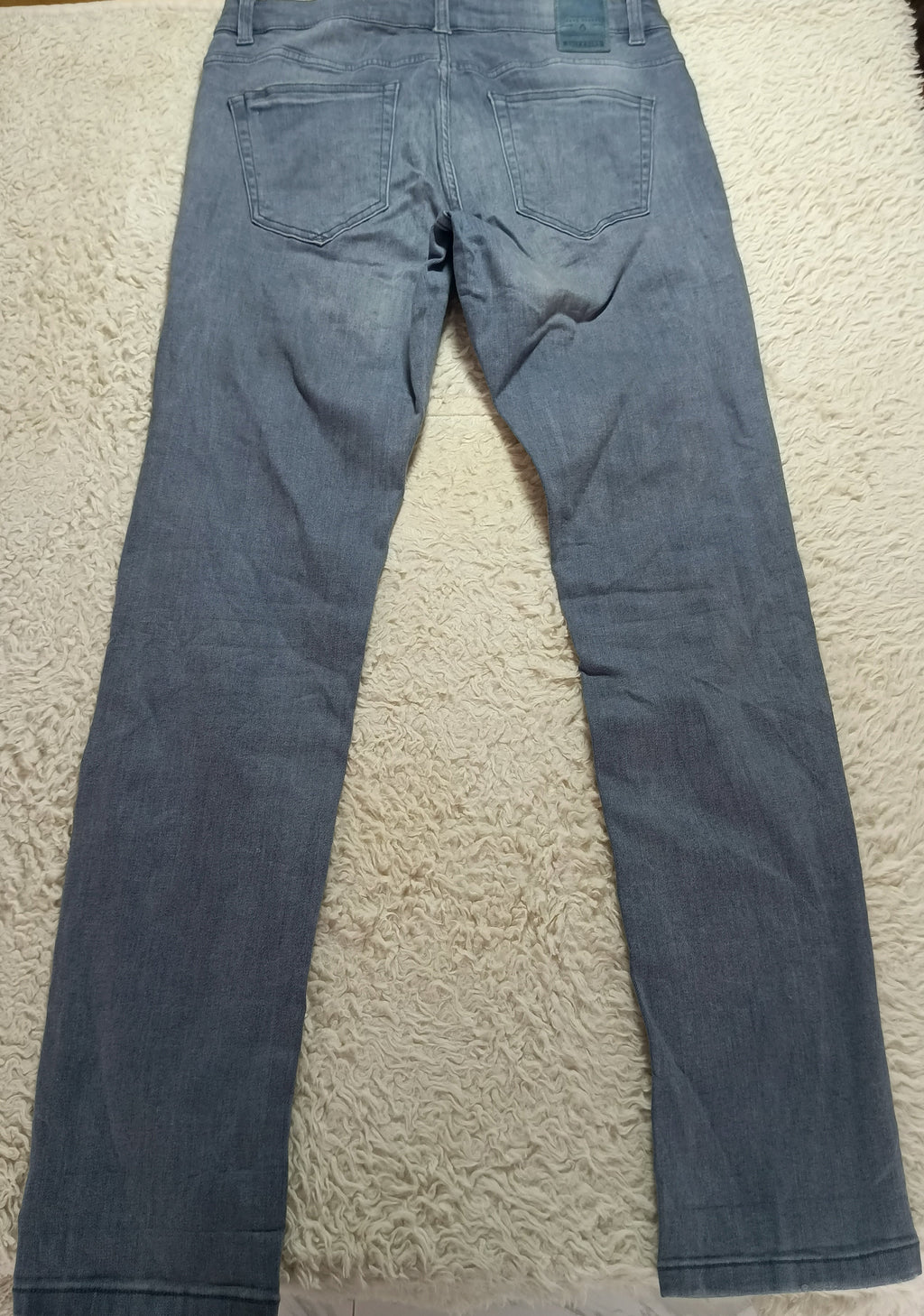 A pair of blue denim jeans laid flat on a white textured rug, showing the back view with the waistband and back pockets visible.