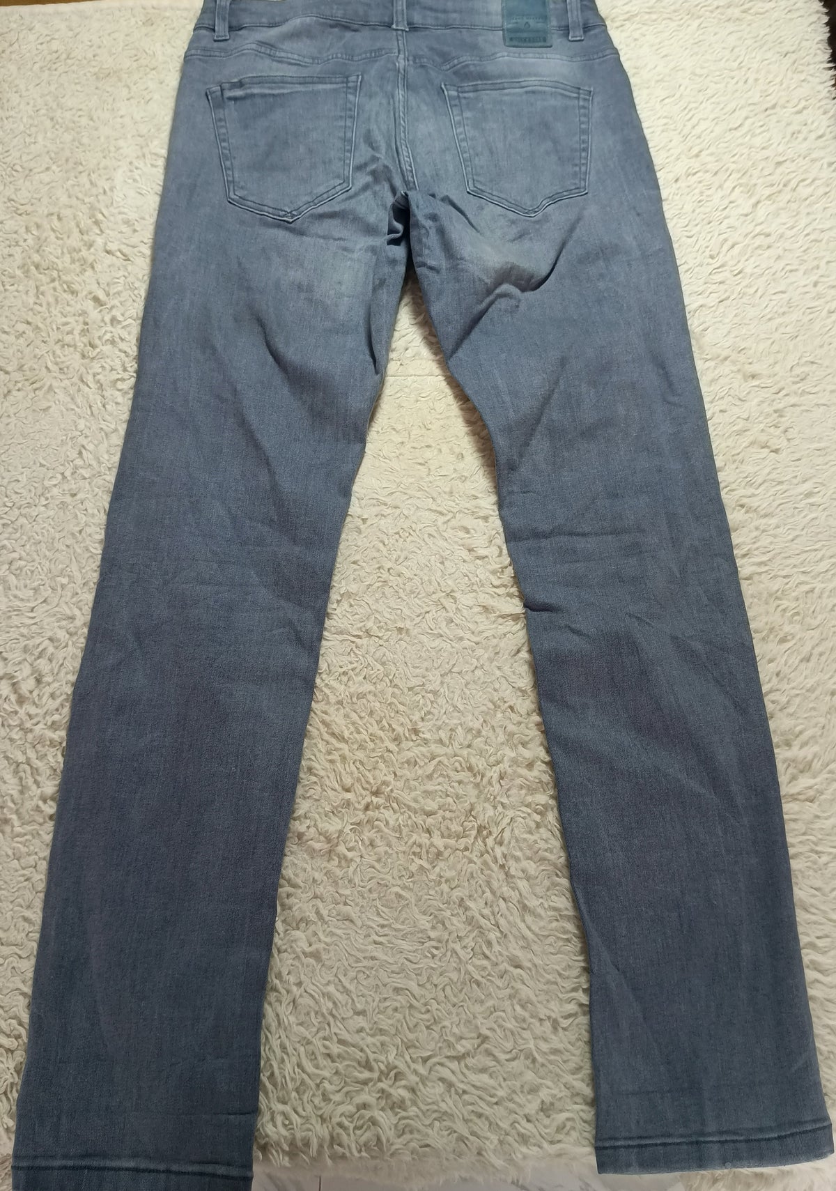 A pair of blue denim jeans laid flat on a white textured rug, showing the back view with the waistband and back pockets visible.