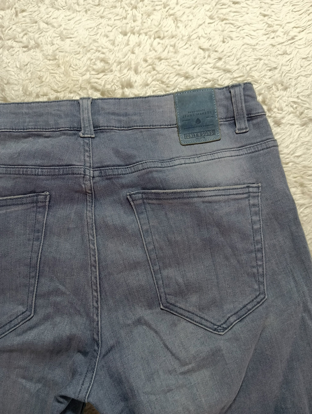 Close-up back view of faded blue denim jeans laid flat on a textured cream rug, showing the waistband, belt loops, and back pockets.
