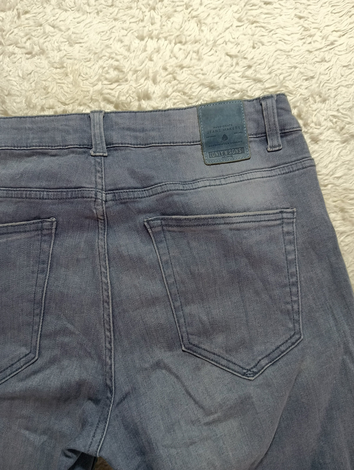 Close-up back view of faded blue denim jeans laid flat on a textured cream rug, showing the waistband, belt loops, and back pockets.