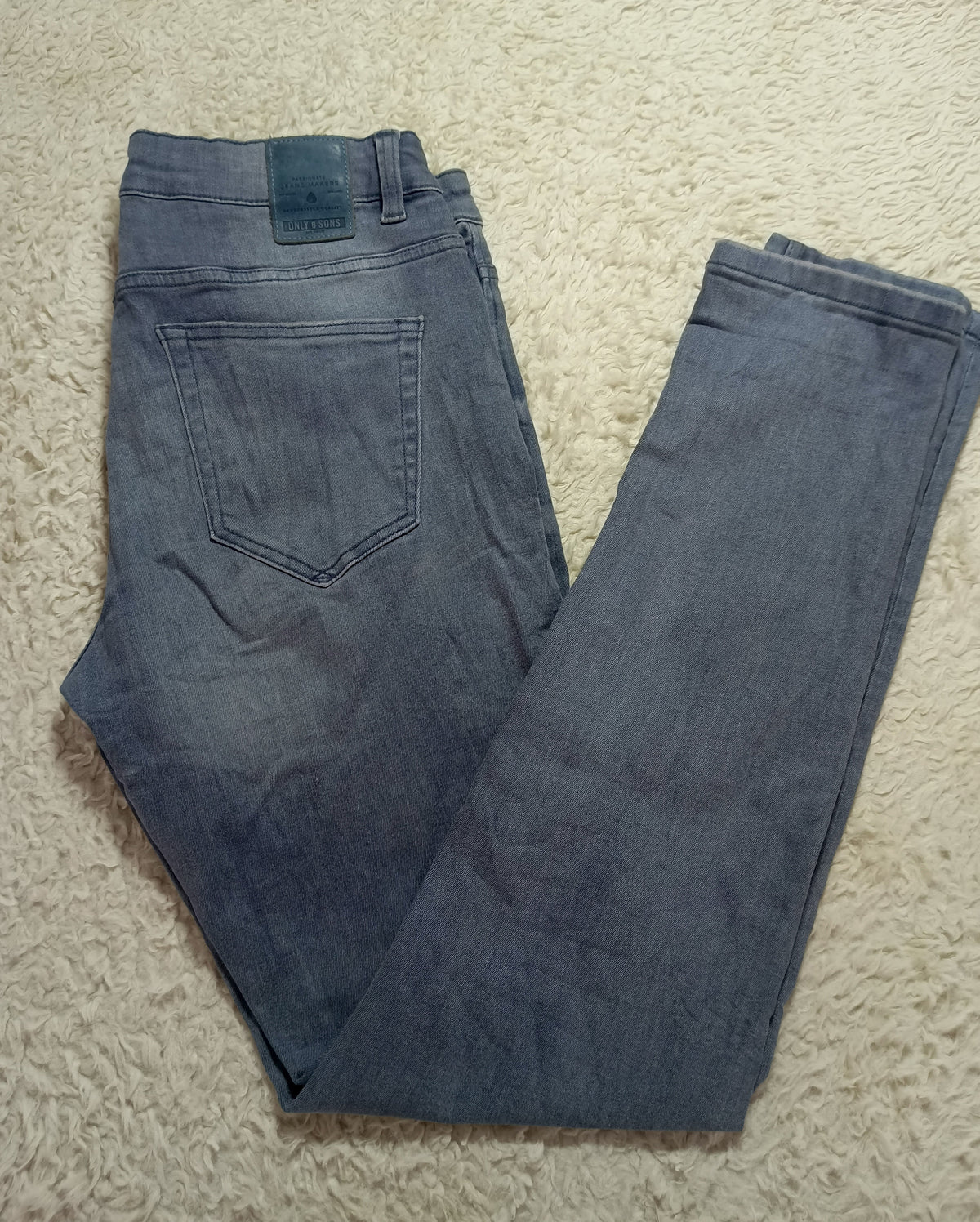 A pair of folded blue denim jeans laid flat on a beige textured rug. The back pocket of the jeans is visible, along with a dark blue leather brand patch near the waistband.