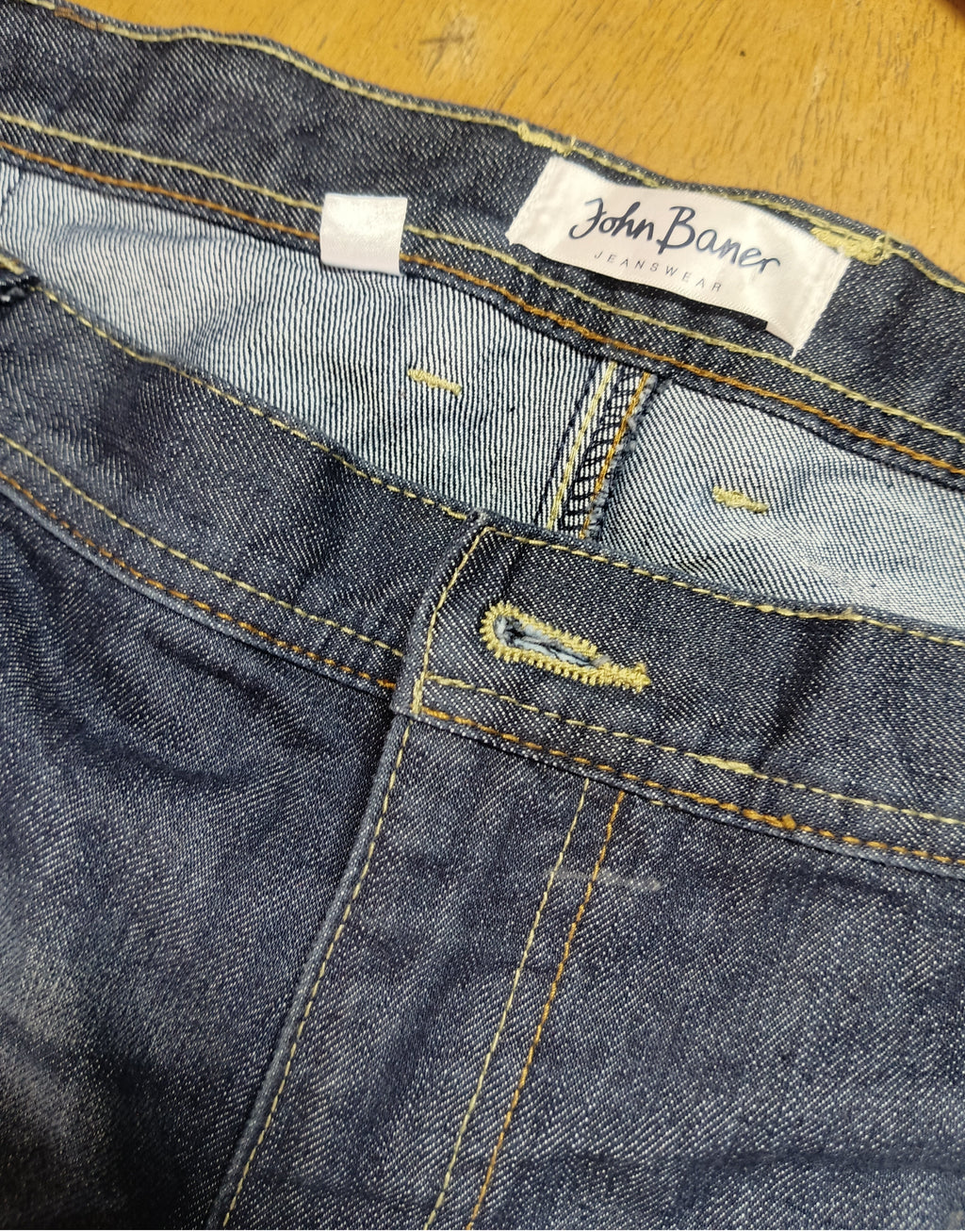 Close-up of a pair of blue denim jeans showing the waistband, inner label reading John Baner, and yellow contrast stitching on a wooden surface.