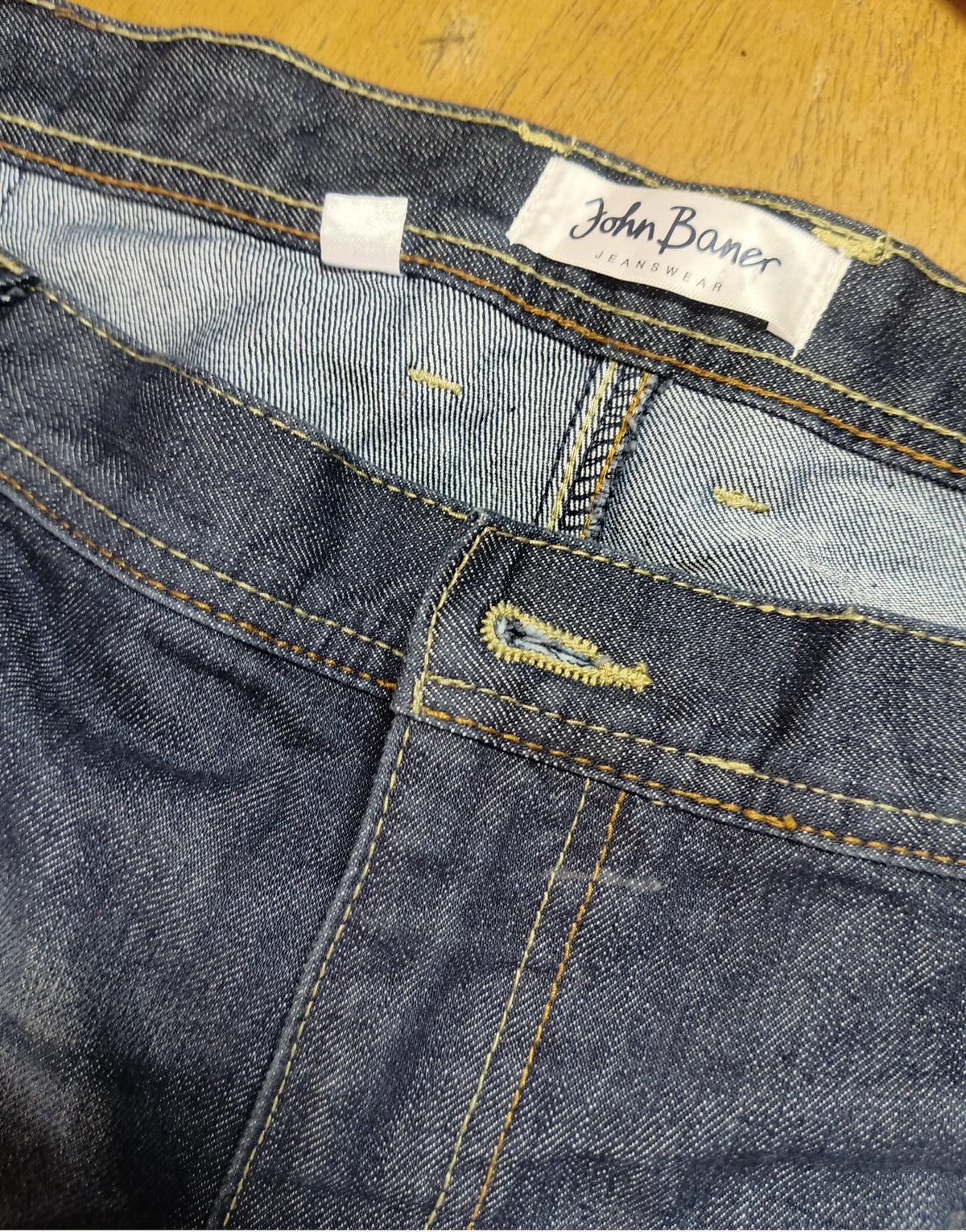 Close-up of a pair of blue denim jeans showing the waistband, inner label reading John Baner, and yellow contrast stitching on a wooden surface.