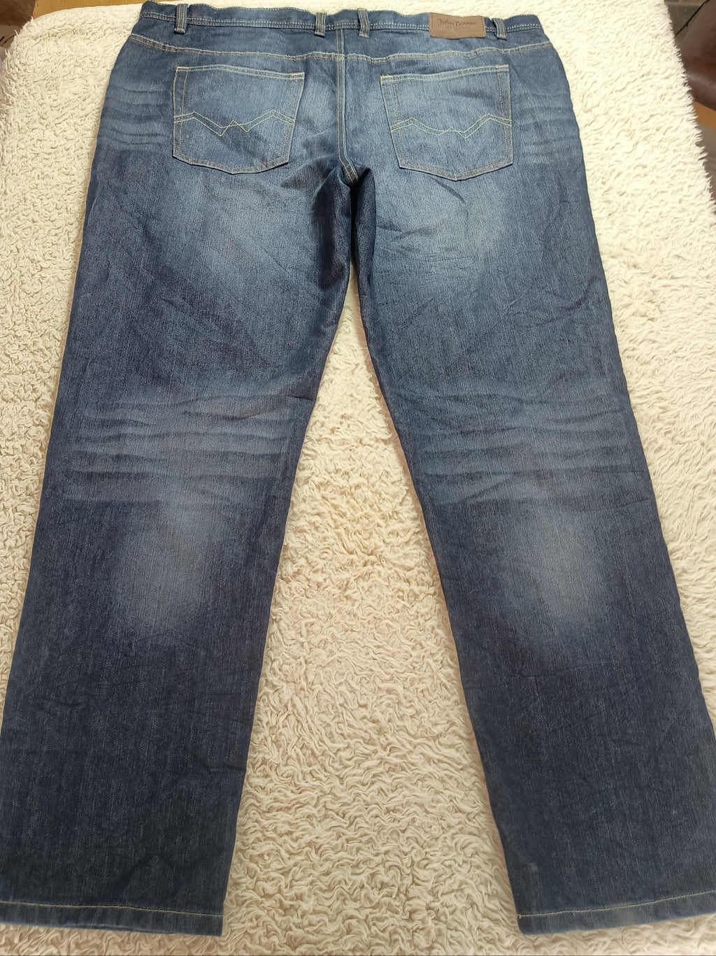 Back view of a pair of blue denim jeans laid flat on a white shag rug, with a wooden table and household items visible in the background.