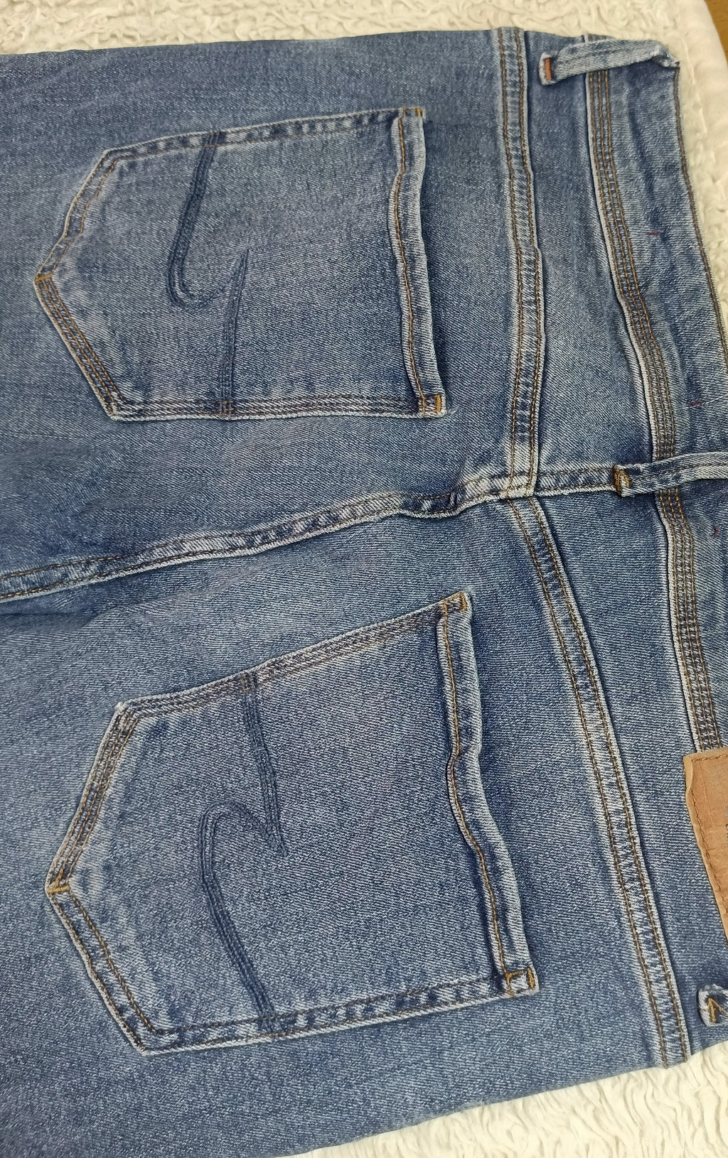 A pair of blue denim jeans laid flat on a textured white blanket, showing the back pockets, waistband, and a brown leather brand patch. The photo is taken with a phone camera, with a wooden floor visible in the top right corner.