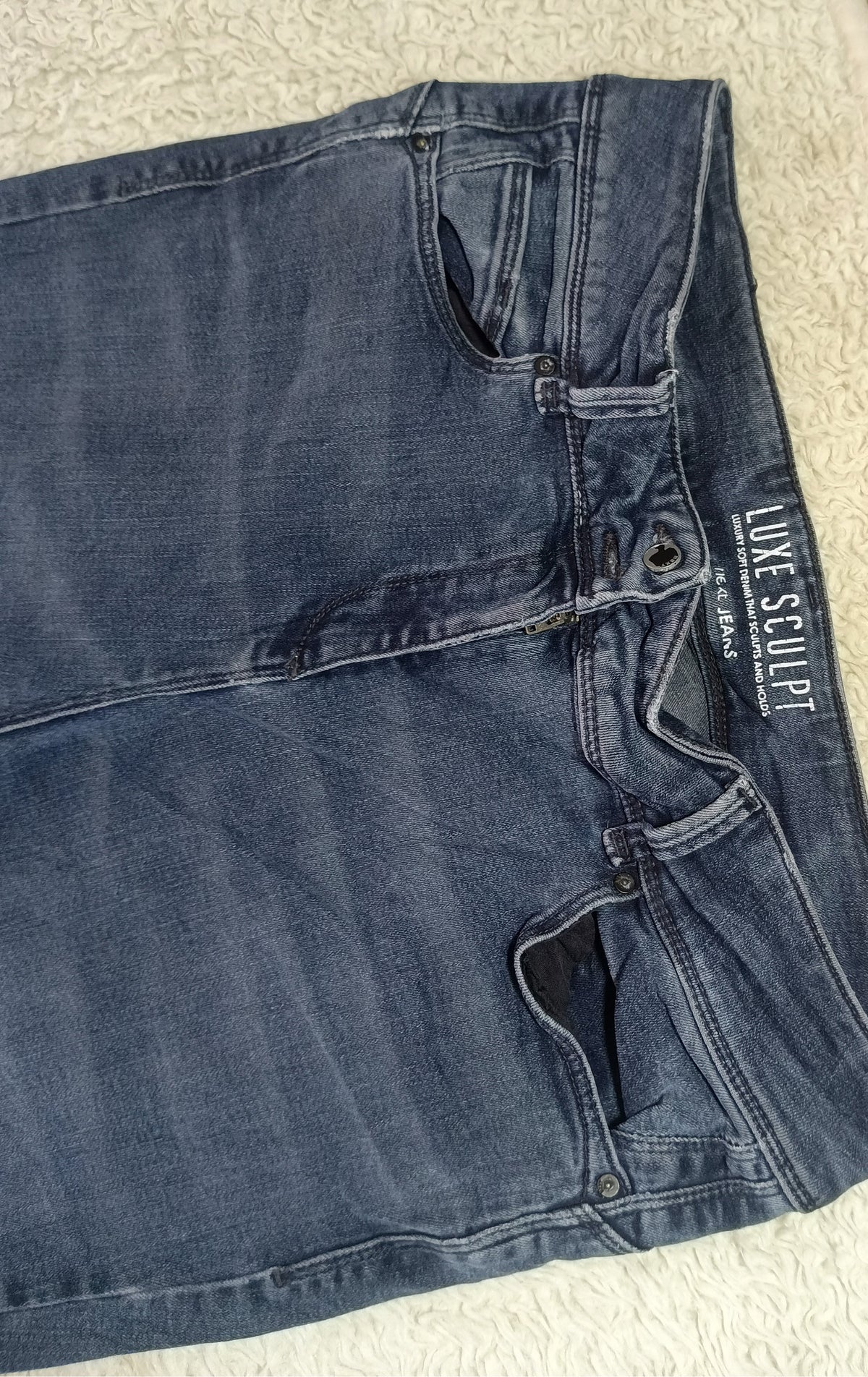 A pair of dark blue denim jeans laid flat on a textured cream fabric surface, showing the waistband, belt loops, front pockets, and a visible 'Luxe Sculpt' brand label.