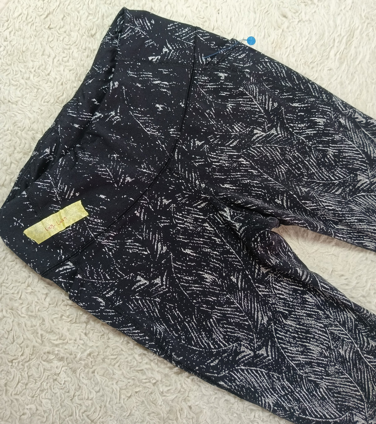 A pair of black leggings with a white abstract brushstroke pattern, laid flat on a white textured rug. A small yellow brand tag is visible on the left leg.