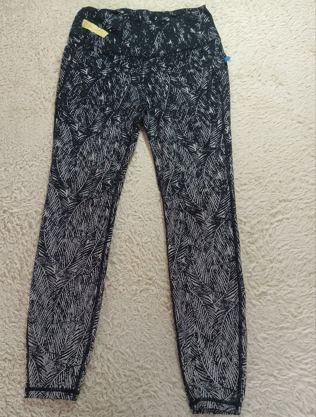 Black and white patterned leggings laid flat on a textured beige carpet. The leggings feature an abstract, leaf-like pattern in black and white, with a small yellow tag near the waistband.