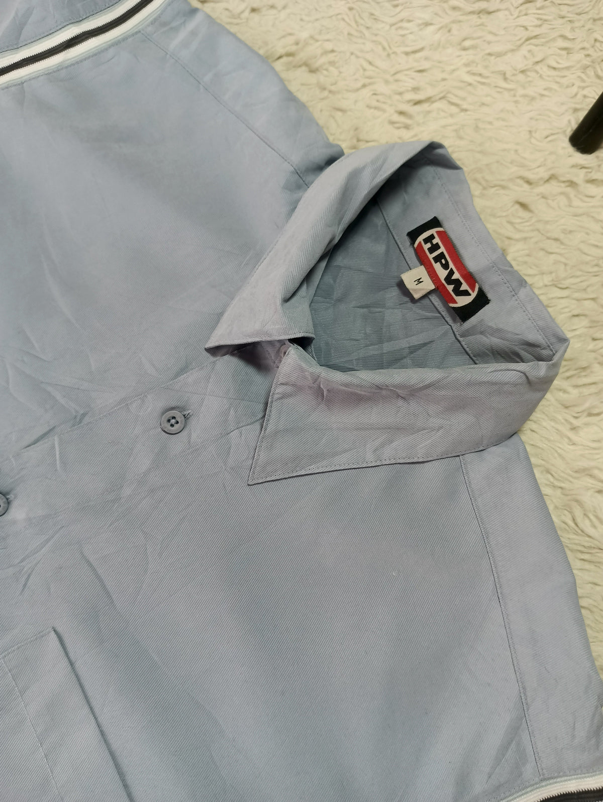 A light blue button-up shirt laid flat on a textured cream rug, with the collar turned up to show a red and black 'HAWK' brand label.