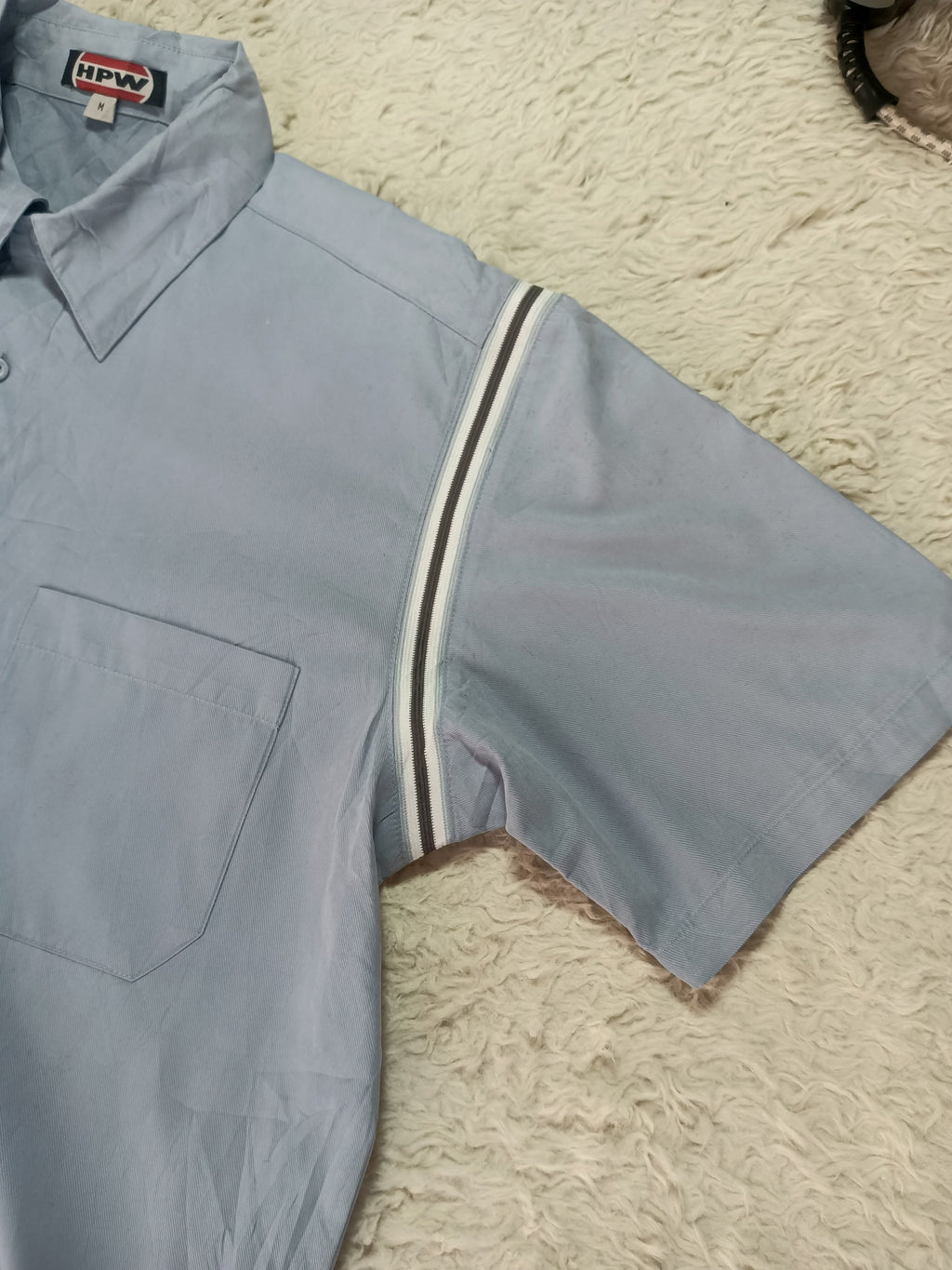A light blue short-sleeve button-up shirt with a striped detail on the sleeve, laid flat on a textured cream rug. The brand label 'HIPPO' is visible on the collar.