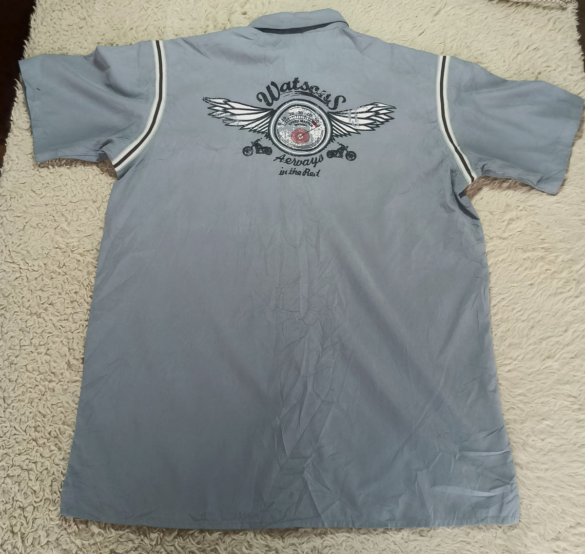 A light blue short-sleeve polo shirt laid flat on a white shag rug, showing the back with a large graphic print featuring wings and the word 'Matsos'. An iron and tripod are visible in the background.